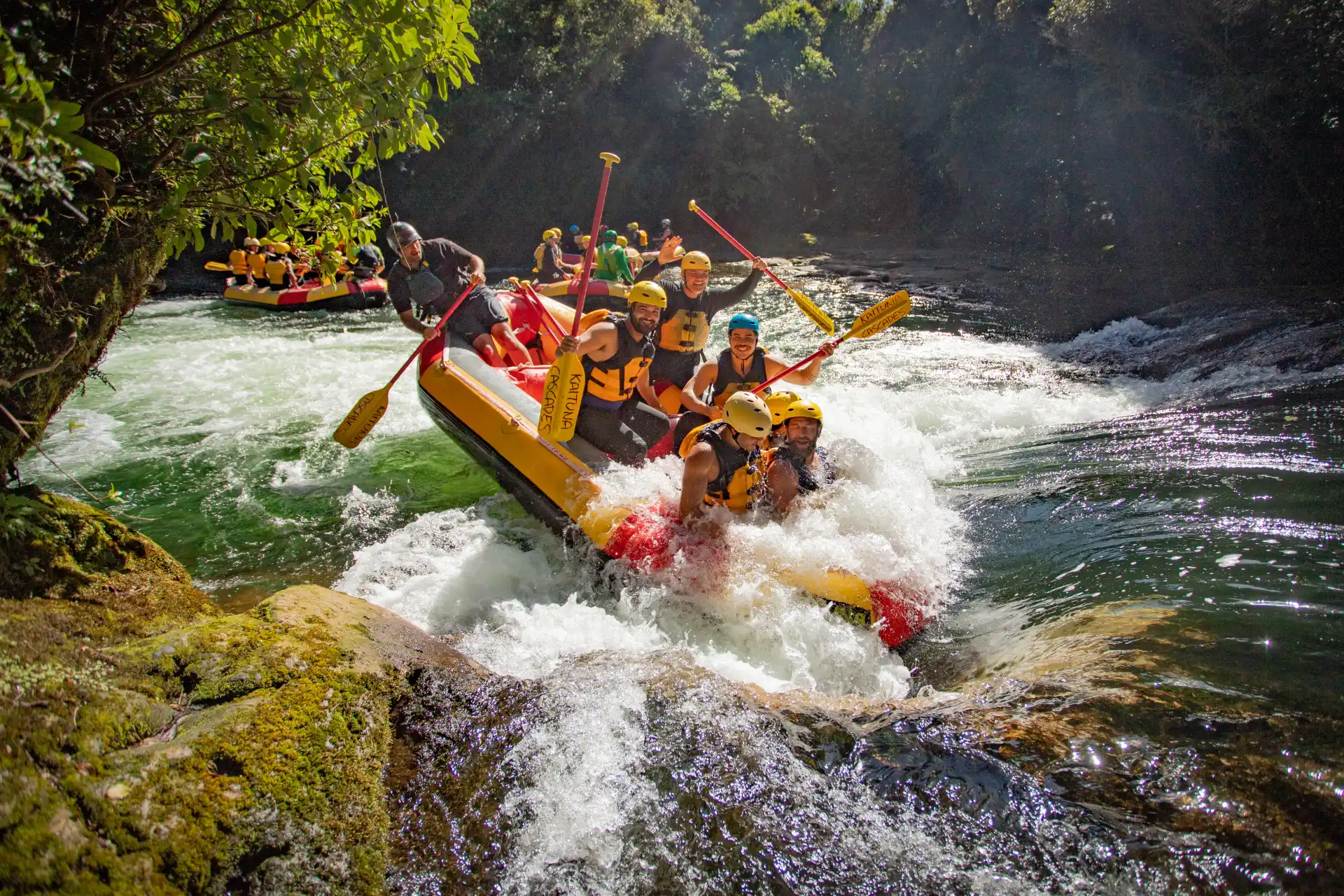 A group of people wearing helmets and life jackets white-water rafting through rapids on a river surrounded by lush green forest.