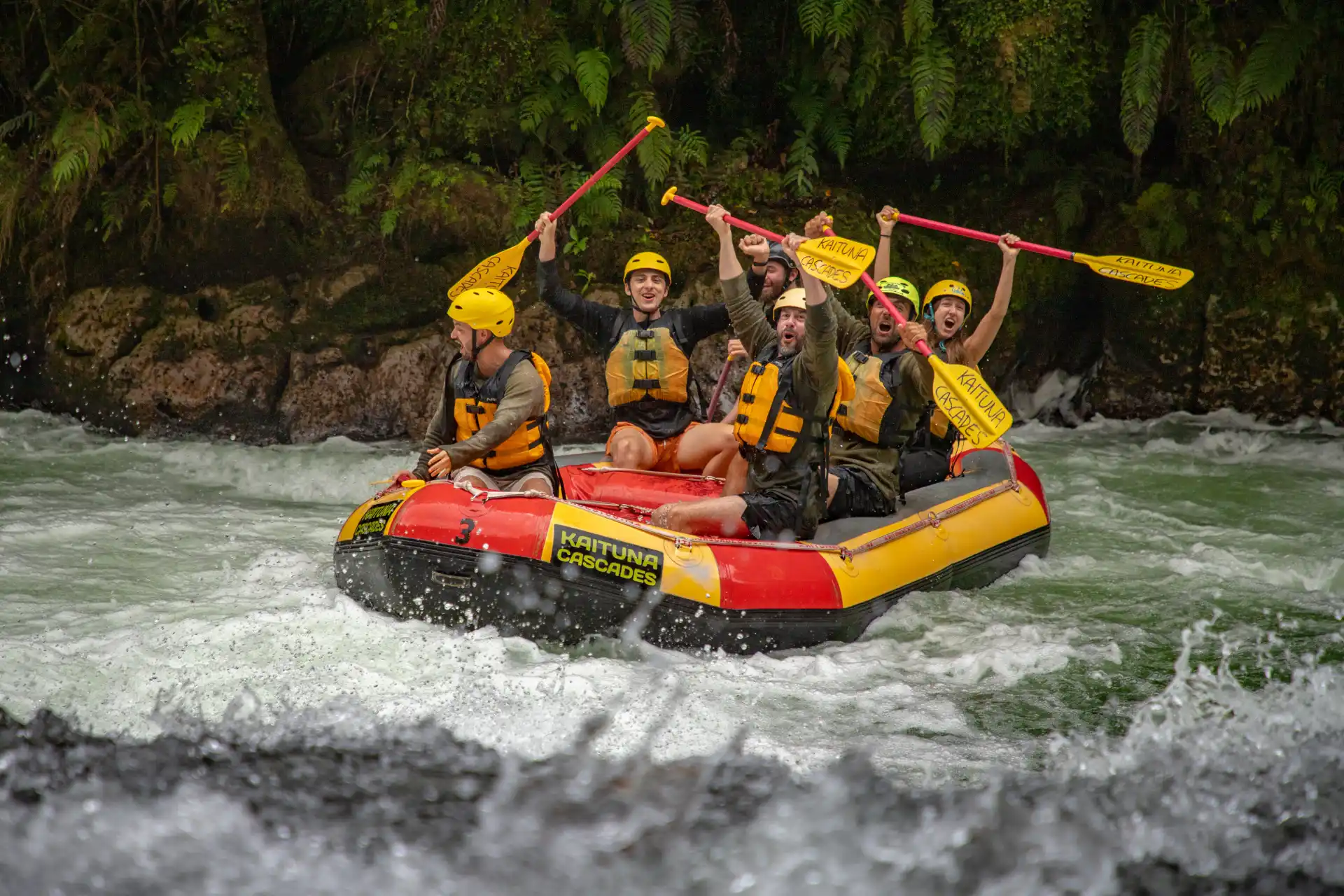 Group celebrating in a Kaituna Cascades rafting boat on white-water rapids surrounded by native forest.